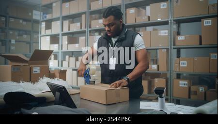 African American Warehouse Clerk Puts Sticker on Cardboard Box with Online Order, Prepares Parcels for Shipping, Scans Code, Uses Tablet Computer. Sorting Center of E-Commerce Store, Delivery Service. Stock Photo
