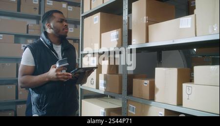 African American Inventory Specialist Walks in Warehouse Retail Facility, Scans Parcels with E-Commerce Online Orders, Checks Delivery Information on Tablet Computer. Sorting Center of Postal Service. Stock Photo