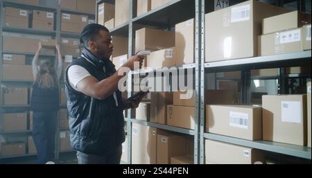 African American Inventory Specialist Walks in Warehouse Retail Facility, Scans Parcels with E-Commerce Online Orders, Checks Delivery Information on Tablet Computer. Sorting Center of Postal Service. Stock Photo