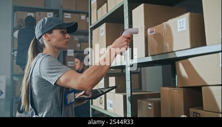 Female Warehouse Worker Scans Cardboard Box with E-Commerce Online Goods on Shelf with Scanner, Using Tablet Computer, Takes Parcel for Shipping to Client. Sorting Center of Delivery Service. Handheld Stock Photo