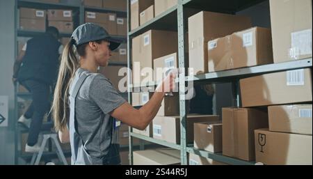 Female Warehouse Worker Scans Cardboard Box with E-Commerce Online Goods on Shelf with Scanner, Using Tablet Computer, Takes Parcel for Shipping to Client. Sorting Center of Delivery Service. Handheld Stock Photo
