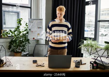 A young man with red hair reviews documents while standing at his desk in a contemporary office. Stock Photo