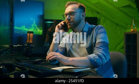 Smiling happy Caucasian broker man guy developer crypto businessman discussing mobile phone at home neon trader male work computer nighttime Stock Photo