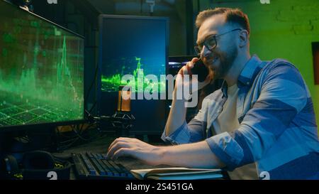 Smiling happy Caucasian broker man guy developer crypto businessman discussing mobile phone at home neon trader male work computer nighttime Stock Photo