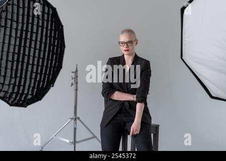 A professional woman poses confidently while seated in a photography studio with softbox lighting equipment, wearing formal business attire. Smiling W Stock Photo