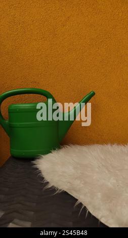Green plastic watering can is leaning on a white fur rug on a yellow background, waiting to be used for gardening Stock Photo