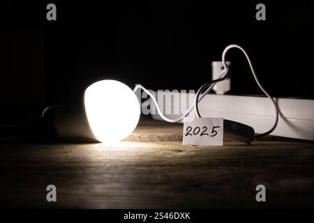 An energy saving light bulb is on a wooden floor near a socket and a card with the number 2025, the light of the lamp and the year 2025 Stock Photo