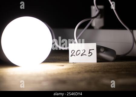 An energy saving light bulb is on a wooden floor near a socket and a card with the number 2025, the light of the lamp and the year 2025 Stock Photo