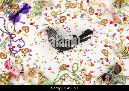 Black mask framed by colourful party decorations such as confetti, pretzels, pearl necklaces and funny glasses on a white background Stock Photo