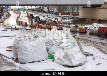 A section of the road under repair with a fence and white bags with paving stones standing on it. Snow on the ground. Stock Photo