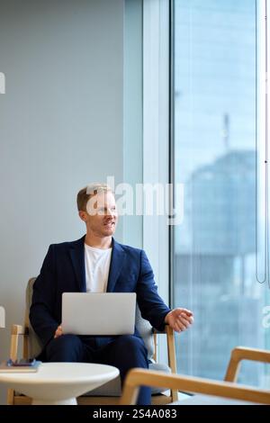 Happy young business man wearing suit thinking of future in office using laptop. Stock Photo