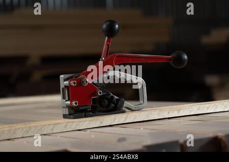 A tie-down tool sits on a stack of wooden planks in the workshop. The tool has a red handle and is designed for connecting tasks. Stock Photo