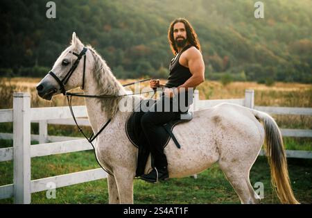 Good looking, hunky cowboy rides horse at beautiful country side nature. Equestrian rides his white well looking horse. Young jockey training his Stock Photo