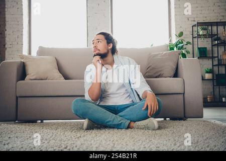 Young man sitting on carpet in modern living room with thoughtful expression Stock Photo
