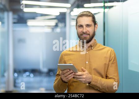 Man standing confidently in a contemporary office while using a tablet. He is smiling and conveys a sense of positive energy, expertise, and a welcoming demeanor ideal for business-related concepts. Stock Photo