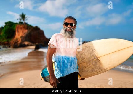 Senior man with surfboard stands on sunny beach. Elderly surfer smiles, wears sunglasses. Active lifestyle, ocean waves background. Enjoy coastal Stock Photo