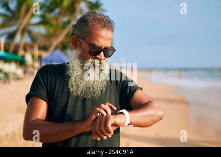 Senior stylish man with gray beard, sunglasses checks fitness tracker on sunny beach. Modern elder enjoys tech gadgets, stays active outdoors. Trendy Stock Photo
