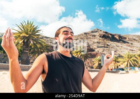 A man is standing on the beach, smiling with closed eyes and doing yoga poses. Stock Photo
