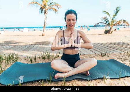A woman is sitting on a mat on the beach, practicing yoga. The scene is peaceful and serene, The woman is focused and relaxed, enjoying the moment Stock Photo