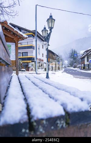 Snow-covered park bench in the centre of a wintry village with traditional houses and street lamps, Enzkloesterle, Calw district, Black Forest, German Stock Photo