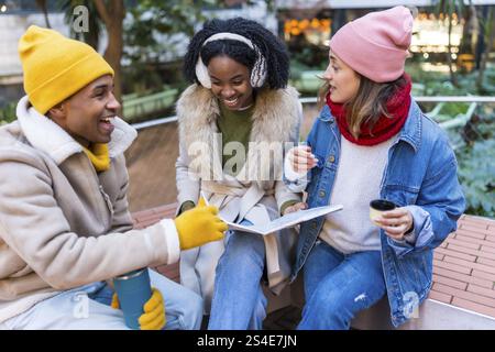Three cheerful young students are sitting on a bench, drinking coffee and studying together on a cold winter day in the city Stock Photo