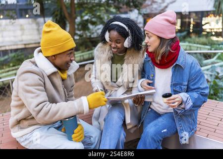 Three happy friends sitting outside in winter, drinking coffee and looking at a map, planning a trip together Stock Photo