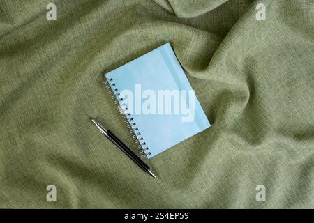 Empty blue hard cover notebook with a ball point pen on olive green fabric backdrop, copy space for text Stock Photo