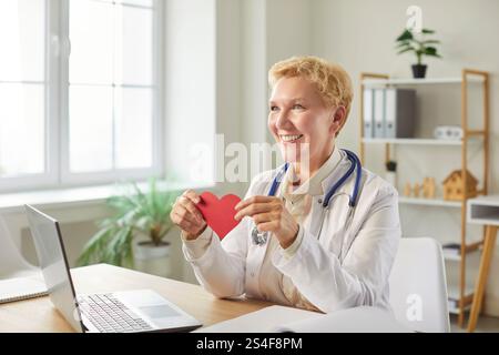Smiling doctor cardiologist holding red heart in hands sitting in medical office Stock Photo