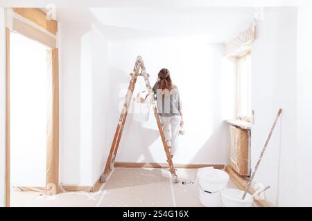 Woman painting home holding a paintbrush in hand stands on a wooden ladder with bucket of paint. Seen from behind in a moment of home decoration, pain Stock Photo