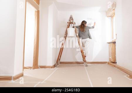 Woman painting home holding a paintbrush in hand stands on a wooden ladder with bucket of paint. Seen from behind in a moment of home decoration, pain Stock Photo