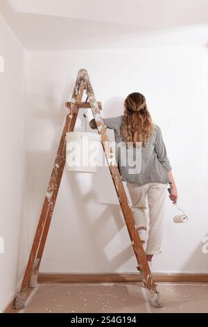 Woman painting home holding a paintbrush in hand stands on a wooden ladder with bucket of paint. Seen from behind in a moment of home decoration, pain Stock Photo