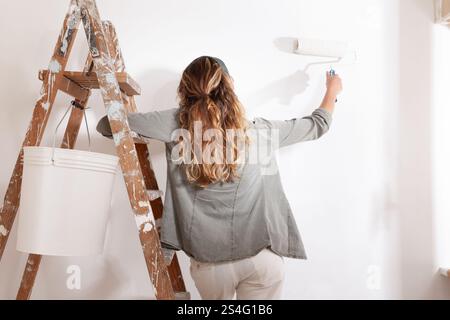 Woman painting home holding a paintbrush in hand stands on a wooden ladder with bucket of paint. Seen from behind in a moment of home decoration, pain Stock Photo