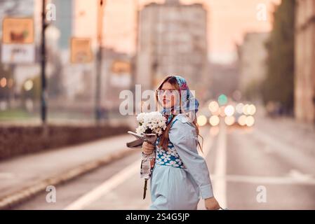 Charm of a woman adorned in an exquisite blue traditional dress, carrying a blue handbag and a bouquet of flowers, gracefully strolling through the ci Stock Photo