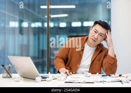 Portrait of disappointed and sad man, businessman dissatisfied looking at camera, near pieces of paper. Asian man doing paperwork inside office. Stock Photo