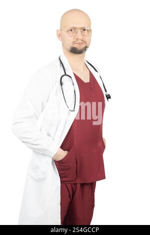 A bald male doctor with glasses wearing red scrubs and a white lab coat poses with hands in pockets. Stock Photo