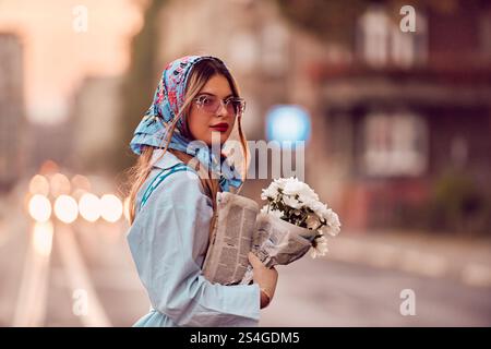 Charm of a woman adorned in an exquisite blue traditional dress, carrying a blue handbag and a bouquet of flowers, gracefully strolling through the ci Stock Photo