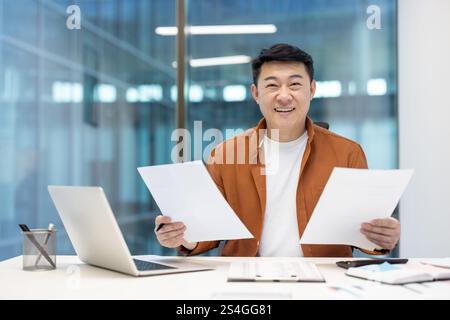 Joyful and successful Asian man doing paperwork inside office, man with documents and reports looking at camera, holding successful investment indicators in hands. Stock Photo