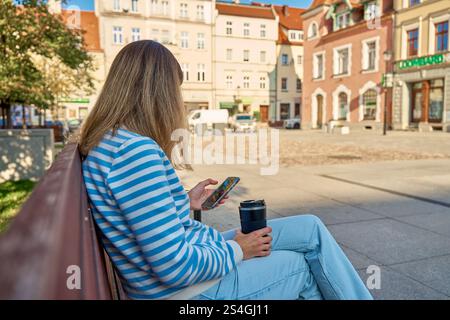 Woman is using smartphone and holding reusable coffee cup while sitting on bench in city park Stock Photo