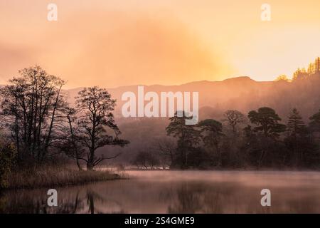 Dramatic atmospheric sunrise landscape at Rydal Water in Lake District during Autumn with moody glow over mist on lake Stock Photo