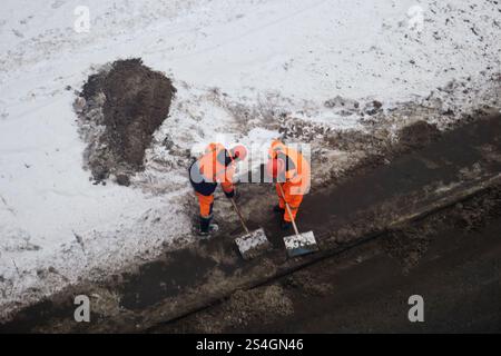 Workers in orange safety suits shoveling snow and dirt on a winter road. Aerial view of winter maintenance work on a snowy street. Stock Photo