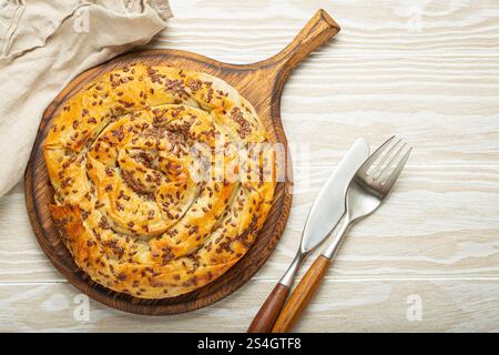 Burek made of filo dough with filling on cutting board, white wooden rustic background top view. Traditional savoury spiral pie of Balkans, Middle Stock Photo