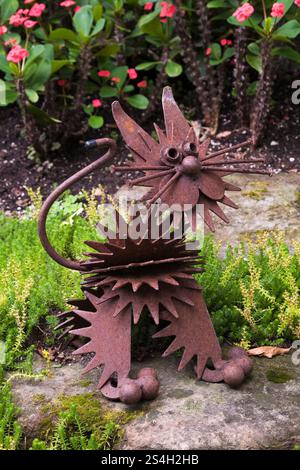 Close-up of rusted metal cat sculpture and red Euphorbia milii 'Crown of Thorns' flowers in background in backyard Zen garden in summer. Stock Photo