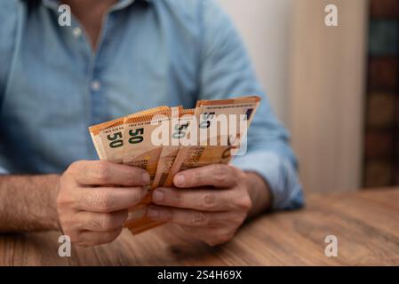 Man holding multiple 50 euro banknotes in both hands on a wooden table. Financial concept highlighting cash handling, savings, and personal finance. Stock Photo