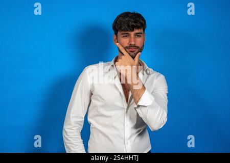 Businessman in white shirt touching his chin, reflecting on important decisions against a vibrant blue backdrop Stock Photo