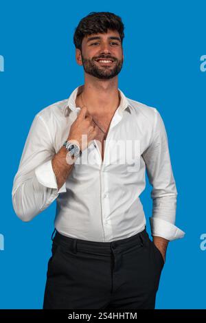 Smiling businessman adjusting his open white shirt collar while posing on a vibrant blue background Stock Photo
