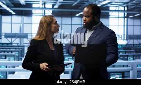 African american admin greeting inspector arriving in data center to supervise operations. IT expert in server hub workspace doing hand shake with supervisor monitoring performance, camera A Stock Photo