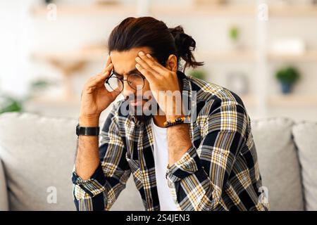 Indian guy sitting on couch at home, touching head Stock Photo