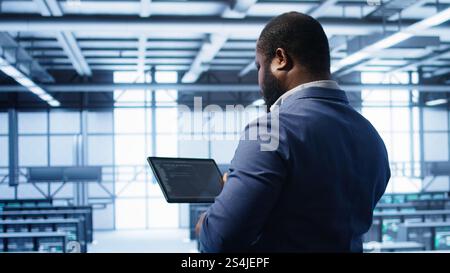IT operator in server room workspace using tablet to patch network issues affecting transfer rates. Engineer in data center fixing errors affecting server rows rigs, camera B Stock Photo
