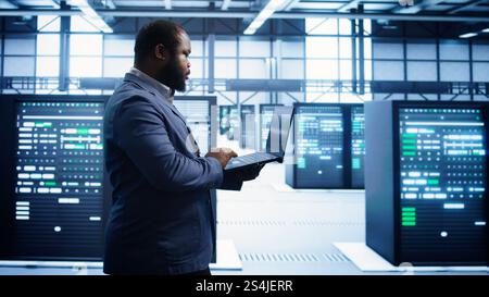 African american man walking in high tech facility, using laptop to patch network issues leading to slow data transfer rates. Data center worker fixing errors affecting supercomputers, camera B Stock Photo