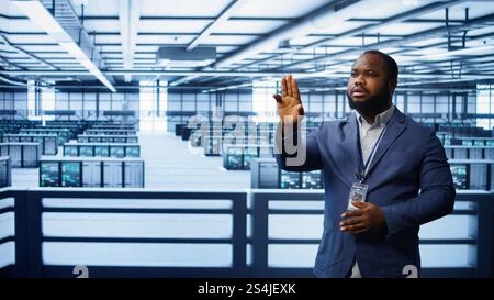 System administrator looking at holograms in data center, monitoring equipment. Employee in server farm interacting with futuristic augmented reality AI visualization, camera B Stock Photo